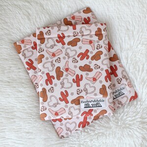 May include: Two pink reusable snack bags with a cowboy boot, cactus, and heart pattern. The bags have a white label with the text "Bocknörticheide" on them.