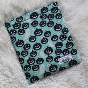 Puede incluir: Una funda de libro de tela azul turquesa con un patrón blanco y negro de calabazas y fantasmas. La funda tiene una pequeña etiqueta blanca con el texto "Buchnordichliebe" en ella.