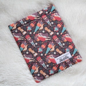 May include: A rectangular book sleeve featuring a repeating pattern of figures in dresses on a dark brown background. The figures are in red, orange, and blue. A white label with the text "Booknerdchickie" is visible.