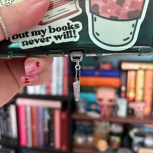 Puede incluir: Una funda transparente para teléfono con un dije. El dije es un cuchillo pequeño con las palabras "Killing it" en la hoja. La funda del teléfono tiene una pegatina que dice "out my books never will!" y una pegatina de una bebida con corazones.