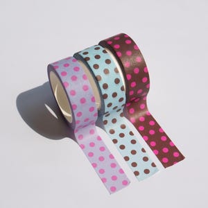 May include: Three rolls of adhesive tape arranged in a row. The first roll is light purple with magenta polka dots, the second is light blue with brown polka dots, and the third is dark brown with magenta polka dots. Each roll has a matching strip extended.