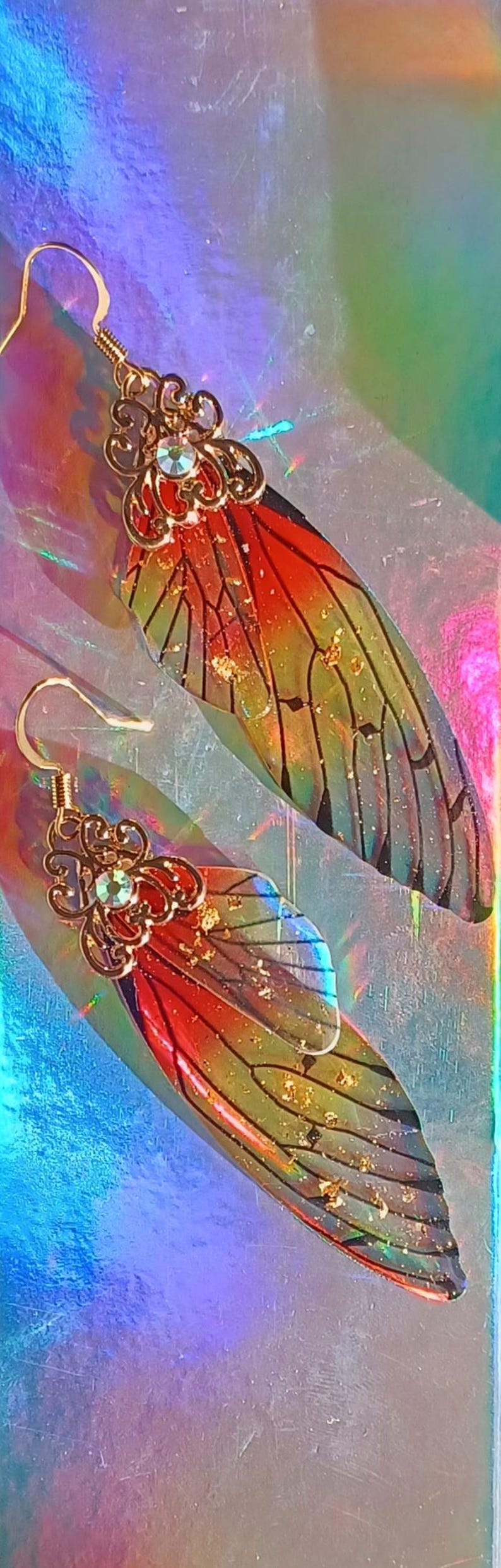 May include: A pair of iridescent butterfly wing earrings. The wings are a gradient of red, orange, yellow, and green with black vein details. Gold-colored filigree and a small gem adorn the top of each wing. Gold-colored ear hooks complete the design.