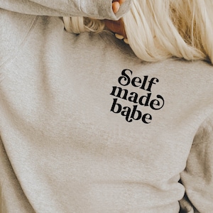 Puede incluir: Una sudadera gris con el texto "Self made babe" en letras negras en la parte delantera.