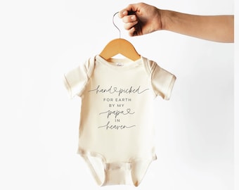 May include: A cream-coloured baby onesie with short sleeves and a black inscription that reads "hand-picked for earth by my papa in heaven".