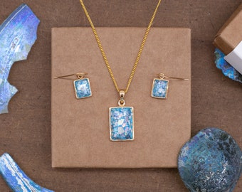 May include: A gold-toned jewelry set featuring a rectangular pendant necklace and matching earrings. The pendant and earrings have a light blue and white mosaic design. The set is displayed on a brown surface.