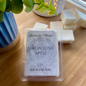 May include: A clear plastic container holding white wax melts, labeled "Macintosh Apple" with "Scents by Nioaka" branding. The container is 4 oz of soy blend wax. Additional wax melts are scattered nearby.