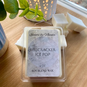May include: A clear plastic container holding a soy blend wax melt. The label reads "Scents by Nioaka" and "Firecracker Ice Pop". The container is 4 oz. and is surrounded by white wax melt cubes. The background includes a white candle holder and greenery.