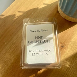 May include: A clear plastic clamshell package containing a soy blend wax melt. The label reads "Scents By Nisaka" and "Pink Grapefruit." The wax melt is 2.5 ounces. The package sits on a wooden surface.