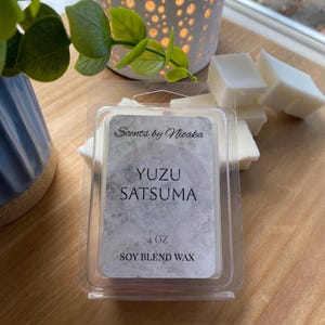 May include: A clear plastic container holding white wax melts, labeled "Yuzu Satsuma" with "Scents by Nioaka" branding. The container holds 4 oz of soy blend wax. Several loose wax melts are next to the container.