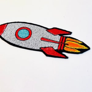 Könnte beinhalten: Ein gestickter Raketen-Patch in Rot, Weiß und Blau mit einer gelben Flamme. Die Rakete fliegt nach rechts.