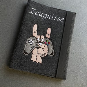 Könnte beinhalten: Ein grauer Filzordner mit einem schwarzen Gummibandverschluss. Der Ordner hat ein gesticktes Design einer Hand, die einen Videospielcontroller hält, mit dem Wort "Zeugnisse" darüber gestickt.