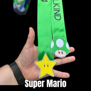 Könnte beinhalten: Ein grünes Band mit der Aufschrift "SCHULKIND 2024" und Verzierungen im Super Mario-Stil. Enthält einen gelben Stern und einen grünen Pilz-Patch. Der Text "Super Mario Schultütenschleife" befindet sich unten.