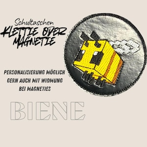 Könnte beinhalten: Ein runder, silberfarbener Aufnäher mit einem gelb-schwarzen, pixeligen Bienen-Design. Der Aufnäher trägt den deutschen Text "Schultaschen Klettie Over Magnetie" und "BIEINE" in einer stilisierten Schriftart. Zusätzlicher Text lautet "Personalisierung möglich gern auch mit Widmung bei Magneties".