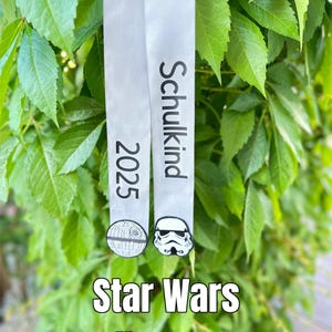 Könnte beinhalten: Zwei weiße Bänder mit den Worten "Schulkind 2025" und Star Wars-Grafiken. Die Bänder hängen vor grünen Blättern. Der Text "Star Wars Schultüten Schleife" befindet sich unten.