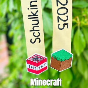 Könnte beinhalten: Zwei Bänder mit Text und Minecraft-Patches. Ein Band sagt "Schulkid" und das andere "2025". Die Patches sind ein TNT-Block und ein Grasblock. Der Text "Minecraft Schultüten Schleife" steht unten.
