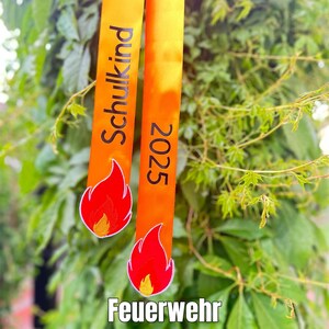 Könnte beinhalten: Zwei orangefarbene Bänder mit den Aufschriften "Schulkind" und "2025", verziert mit gestickten Flammenmotiven. Die Bänder sind vor einem Hintergrund aus grünem Laub angeordnet, mit dem Text "Feuerwehr Schultüten Schleife" am unteren Rand.