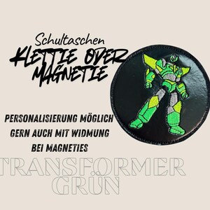 Könnte beinhalten: Ein runder, schwarzer Aufnäher mit einem grünen und silbernen Roboter-Design. Der Aufnäher hat den Text "Schultaschen KLETTE OVER MAGNETIE" und "TRANSFORMER GRUN". Zusätzlicher Text lautet "PERSONALISIERUNG MÖGLICH GERN AUCH MIT WIDMUNG BEI MAGNETIES".
