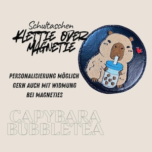 Könnte beinhalten: Ein runder, marineblauer Aufnäher mit einem Cartoon-Wasserschwein, das Bubble Tea trinkt. Das Wasserschwein ist hellbraun und hält einen blau-weißen Bubble Tea-Becher. Der Text "CAPYBARA BUBBLETEA" ist sichtbar.
