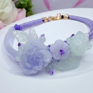 May include: A lavender bracelet with floral and leaf accents. The bracelet features a woven cord and a gold clasp. The decorative elements include a large carved flower, smaller flowers, and leaves in shades of lavender and green.