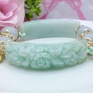 May include: A light green jade bracelet with carved floral details. The bracelet is composed of two curved jade pieces connected by beaded sections with gold accents. The jade has a translucent quality, and the bracelet is displayed on a white surface.