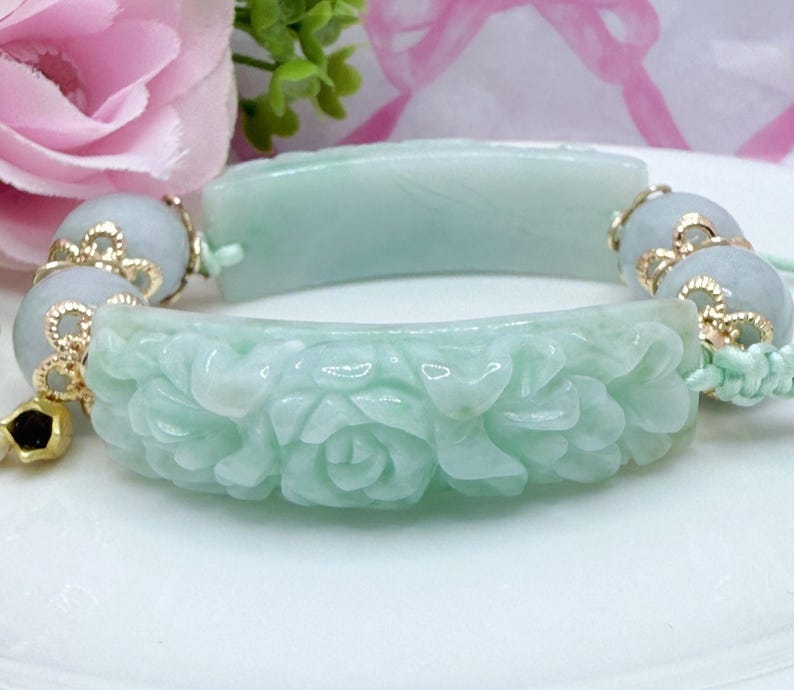 May include: A light green jade bracelet with carved floral details. The bracelet is composed of two curved jade pieces connected by beaded sections with gold accents. The jade has a translucent quality, and the bracelet is displayed on a white surface.