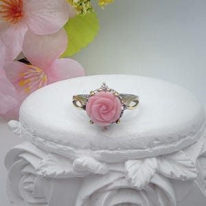 Peut inclure: Une bague argentée avec une pierre centrale sculptée en forme de rose rose. La rose est entourée d'un halo de petites pierres claires et scintillantes serties dans une monture métallique. L'anneau présente un design fendu avec des accents dorés.