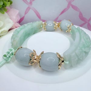 May include: A jade bracelet featuring carved green leaf accents and light blue beads. The bracelet is strung with a light green cord and has gold-colored decorative elements. The bracelet is displayed on a white surface.