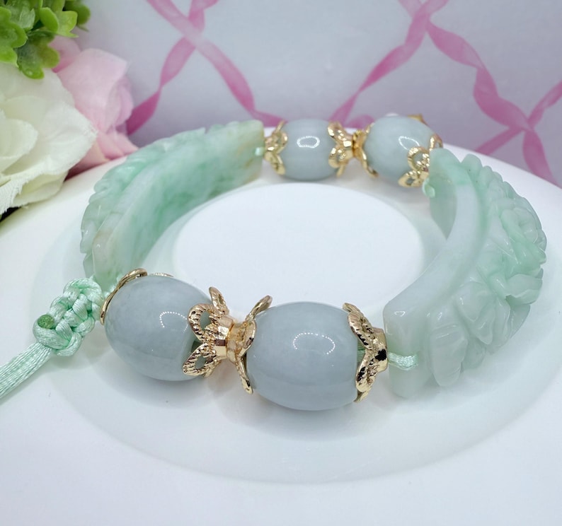 May include: A jade bracelet featuring carved green leaf accents and light blue beads. The bracelet is strung with a light green cord and has gold-colored decorative elements. The bracelet is displayed on a white surface.