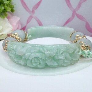 May include: A light green jade bracelet with carved floral details. The bracelet features gold accents and light green beads. The main section of the bracelet is carved with a floral pattern. The bracelet is on a white surface.