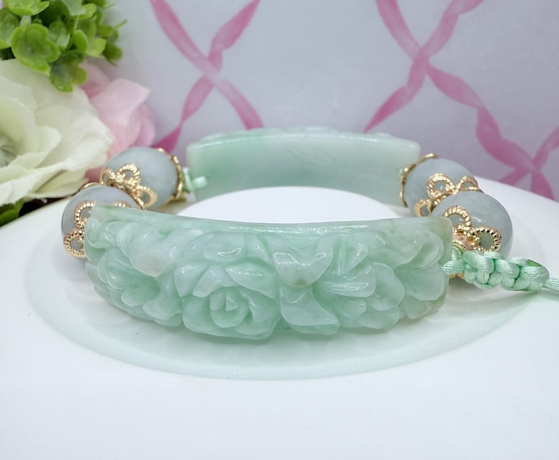 May include: A light green jade bracelet with carved floral details. The bracelet features gold accents and light green beads. The main section of the bracelet is carved with a floral pattern. The bracelet is on a white surface.
