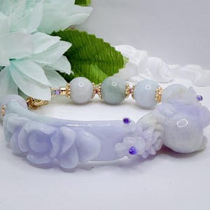 May include: A jade bracelet featuring a carved floral design in shades of lavender and white. The bracelet includes gold accents and round jade beads in varying shades of green and white. Small purple gemstones are also incorporated into the design.