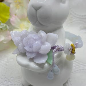May include: A lavender jade flower charm bracelet with a purple cord and gold accents. The bracelet features a carved jade flower charm and several smaller jade beads.
