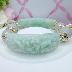 May include: A light green jade bracelet with carved floral details. The bracelet features a central carved section and is accented with gold-colored beads and connectors. The jade has a translucent quality, and the bracelet is displayed on a white surface.