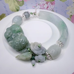 May include: A jade bracelet with a curved, light green section and various jade beads and carvings. The bracelet features a detailed carving of a creature, along with floral and rounded jade elements. Silver-colored metal accents connect the jade pieces.