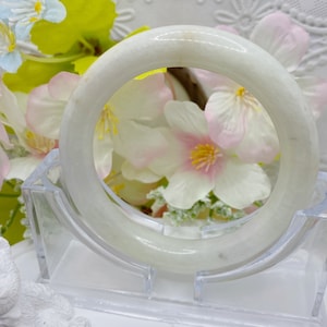 May include: A white jade bangle bracelet with a smooth, polished finish. The bangle is displayed on a clear plastic stand.