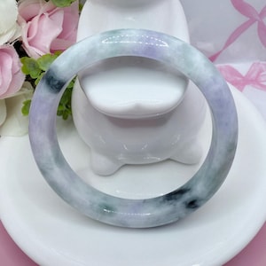 May include: A jade bangle bracelet with a smooth, polished surface. The bracelet features a blend of colors, including lavender, green, and black, creating a marbled effect. The bangle is circular and appears to be a solid piece of stone.
