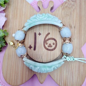 May include: A jade bracelet with a light green floral carved design. The bracelet features light blue oval beads with gold accents. The number "16" is visible on the wooden background, along with a small cartoon face.