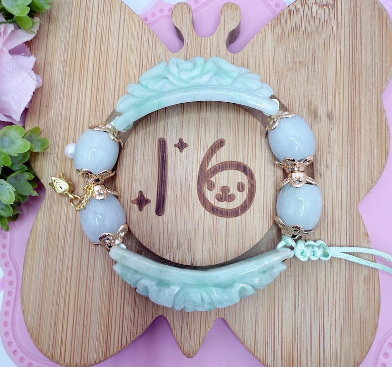 May include: A jade bracelet with a light green floral carved design. The bracelet features light blue oval beads with gold accents. The number "16" is visible on the wooden background, along with a small cartoon face.