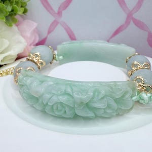 May include: A jade bracelet with carved floral and geometric designs. The bracelet features light green jade segments with gold-colored accents and round jade beads. The bracelet is displayed on a white surface with a floral background.