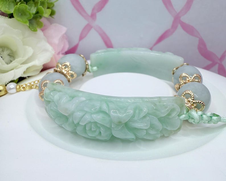 May include: A jade bracelet with carved floral and geometric designs. The bracelet features light green jade segments with gold-colored accents and round jade beads. The bracelet is displayed on a white surface with a floral background.