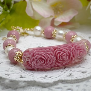 May include: A pink and white bracelet with carved rose details. The bracelet is made of pink and white beads and a carved pink rose charm.