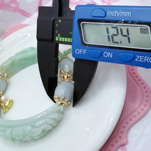 May include: A light green jade bracelet with gold accents is measured with a blue digital caliper. The bracelet features carved floral and oval-shaped jade beads. The caliper displays a measurement of 12.4 mm.
