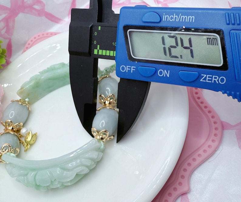 May include: A light green jade bracelet with gold accents is measured with a blue digital caliper. The bracelet features carved floral and oval-shaped jade beads. The caliper displays a measurement of 12.4 mm.