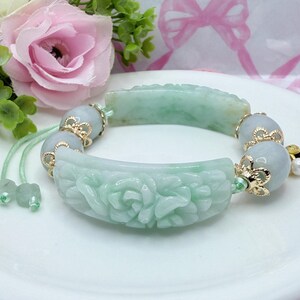 May include: A jade bracelet with carved floral designs and gold accents. The bracelet features light green jade pieces and round beads, strung together with a green cord. A pearl charm hangs from the bracelet.