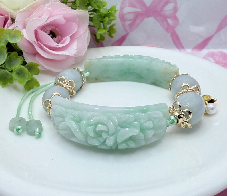 May include: A jade bracelet with carved floral designs and gold accents. The bracelet features light green jade pieces and round beads, strung together with a green cord. A pearl charm hangs from the bracelet.