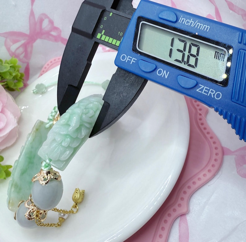 May include: A light green jade pendant with carved floral details is measured with a digital caliper. The measurement reads 13.8 mm. The pendant is attached to a gold chain with gray beads and a gold floral charm. The pendant rests on a white plate.