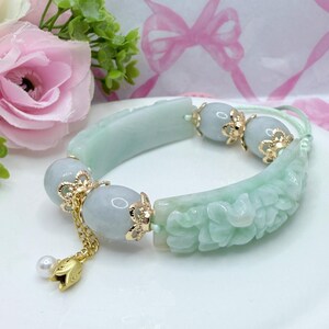 May include: A light green jade bracelet with carved floral details and gold accents. The bracelet features three light blue beads and a gold charm with a pearl. The bracelet is on a white surface.
