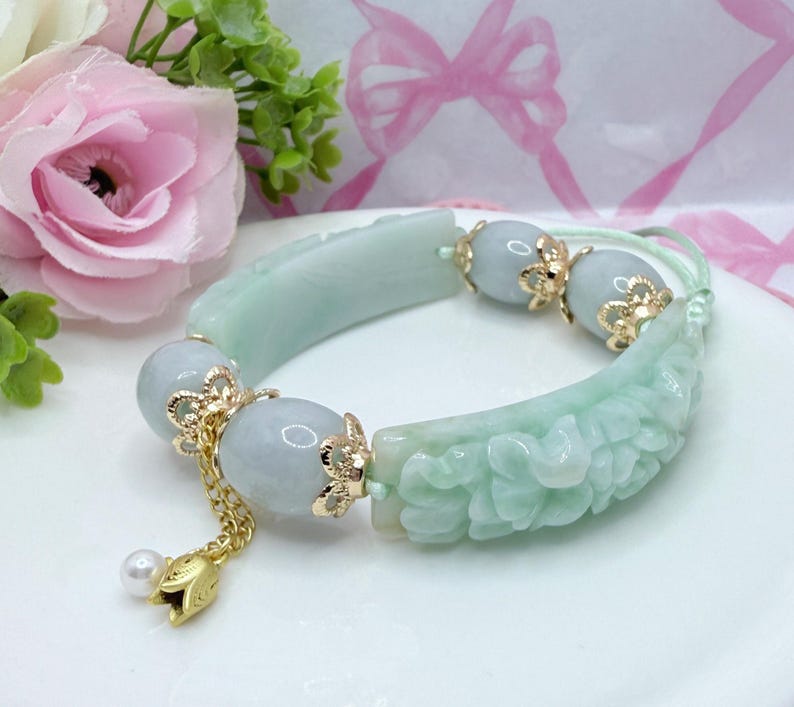 May include: A light green jade bracelet with carved floral details and gold accents. The bracelet features three light blue beads and a gold charm with a pearl. The bracelet is on a white surface.
