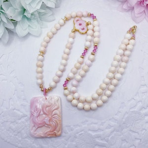 May include: A pink and white coral necklace with a pink coral pendant featuring a carved fox design. The necklace is made with small round beads and has gold accents.