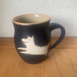 May include: A black ceramic mug with a white silhouette of a dog jumping. The mug has a beige rim.